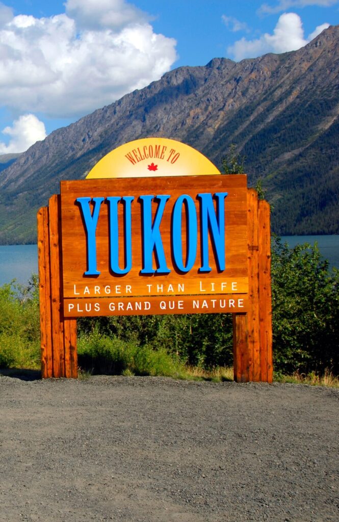 Pictured is a wooden sign in nature near a lake and mountain that has text reading " Welcome to Yukon. Larger than life. Plus grand que nature"